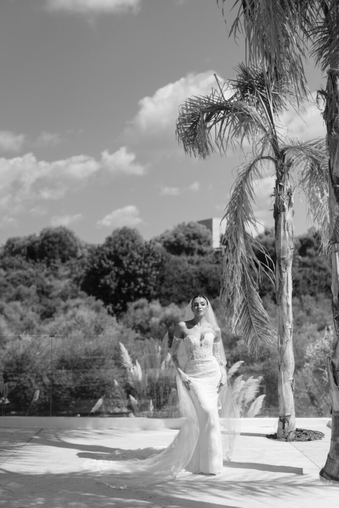 A bride in a fitted wedding dress and veil stands on a sunlit patio near palm trees, with lush greenery and a partly cloudy sky in the background. The photo is in black and white.