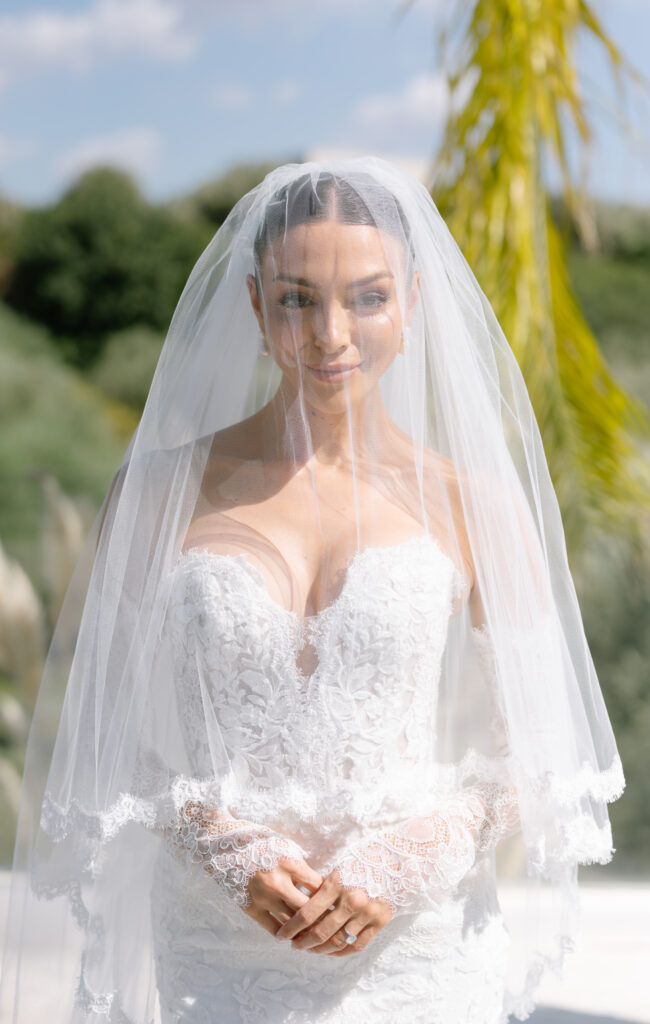 A bride in a white lace strapless wedding dress stands outdoors, smiling softly with her eyes closed under a sheer veil. Greenery and blue sky are visible in the sunny background.