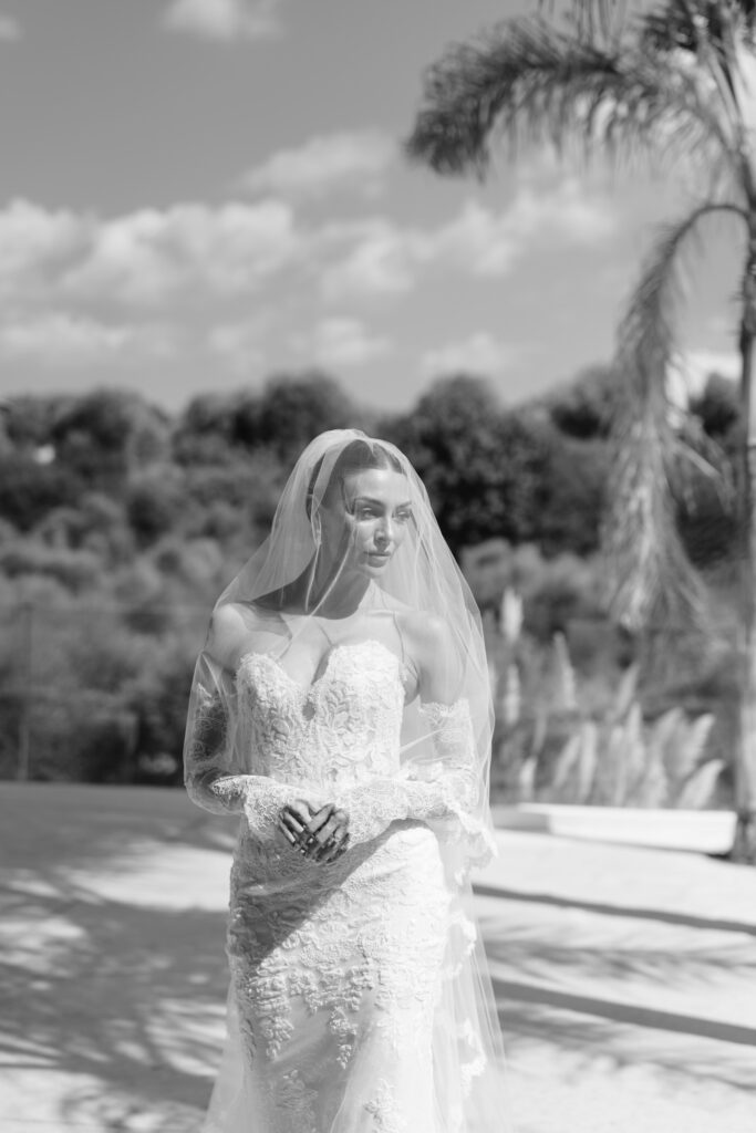 A bride in a lace wedding dress and veil stands outdoors in sunlight, with trees and a palm tree in the background. The image is in black and white.