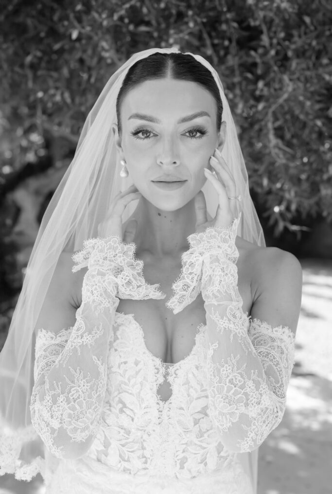 A bride in a lacy, off-the-shoulder wedding dress and veil stands outdoors, looking directly at the camera with her hands gently touching her face.