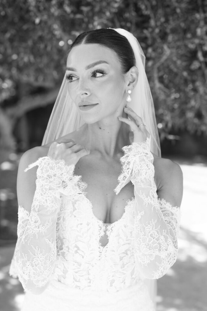 A bride in a lace wedding dress and veil poses outdoors, looking to the side with one hand touching her neck, the other near her chin. The background is softly blurred with trees. The photo is in black and white.