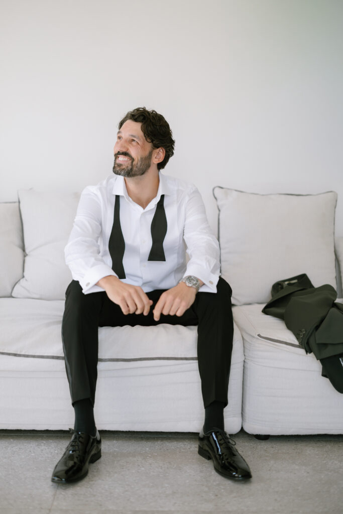 A man with a beard sits on a light-colored couch, smiling and looking to the side. He wears a white dress shirt with a loosened black bow tie around his neck, black pants, and shiny black shoes. A suit jacket rests beside him.