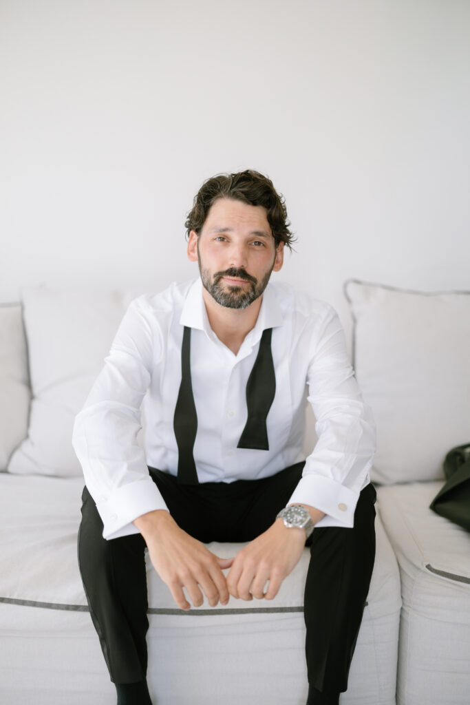 A man with wavy dark hair and a beard sits on a light-colored sofa, wearing a white dress shirt with an untied black bow tie around his neck, and black pants. He looks calmly at the camera.