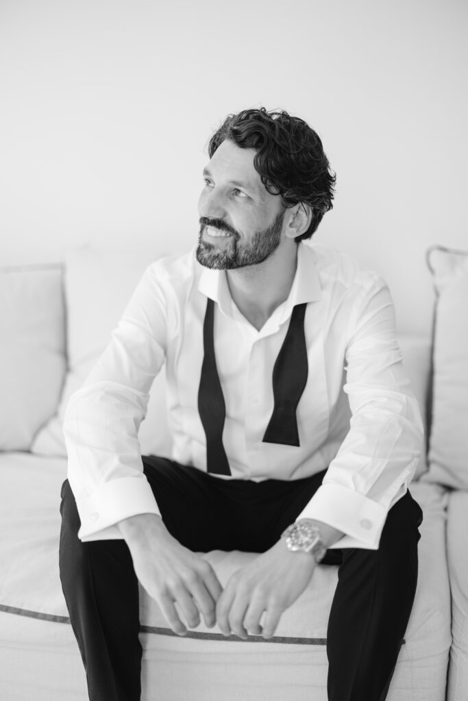 A man with a beard and wavy hair sits on a couch, smiling and looking to the side. He wears a white dress shirt with an undone black bow tie and black trousers. The image is in black and white.
