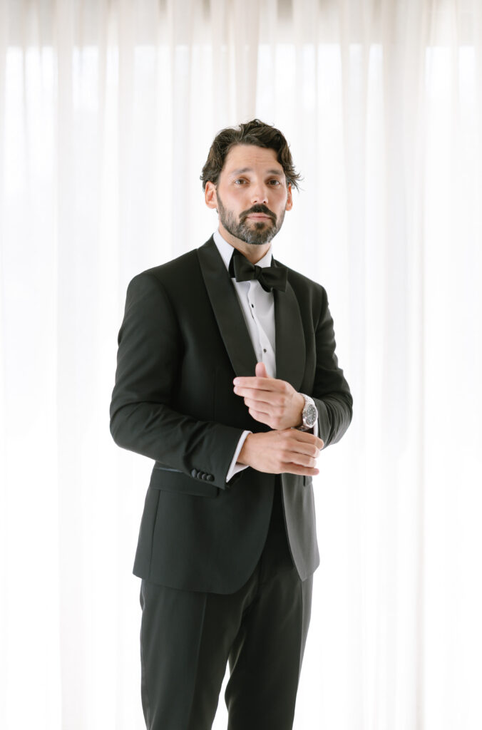 A man with dark hair and a beard wears a black tuxedo with a bow tie, standing in front of sheer white curtains. He adjusts his sleeve and looks confidently at the camera.