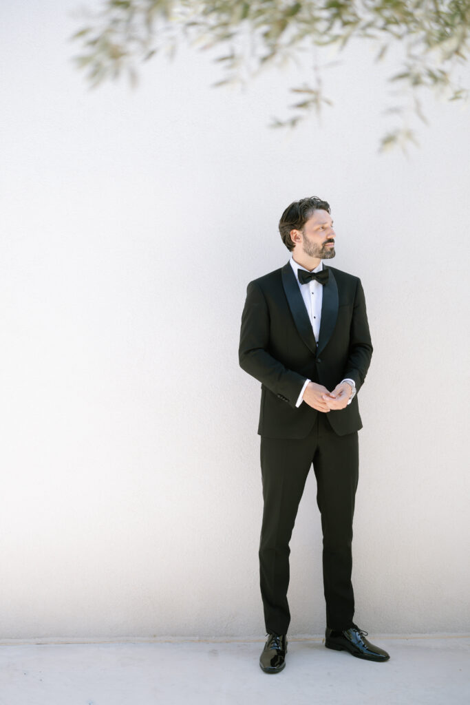 A man in a black tuxedo with a bow tie stands against a plain light-colored wall, looking to his left. There are blurred green leaves in the upper right corner of the image.