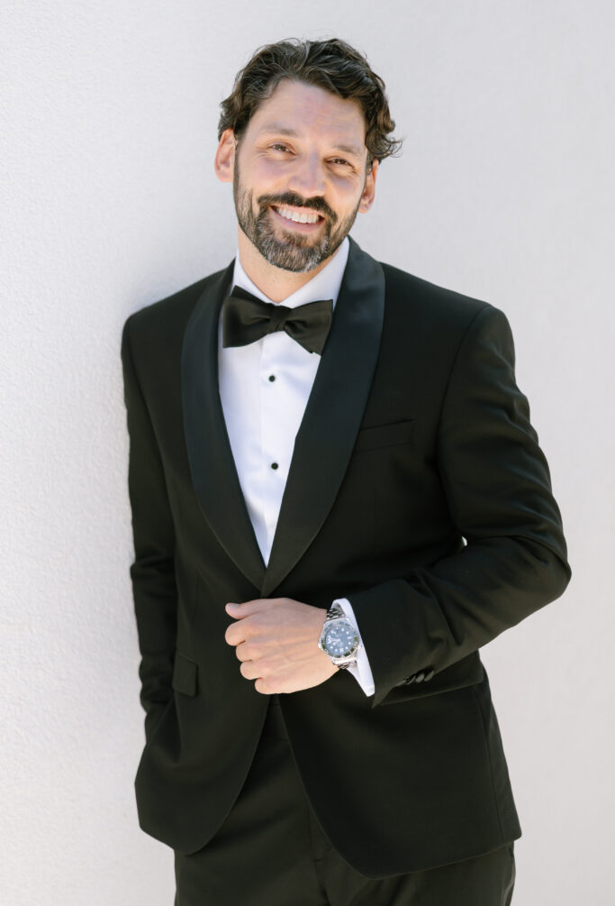 A man with wavy dark hair and a beard, wearing a black tuxedo with a bow tie and a watch, stands against a light-colored wall, smiling confidently at the camera.