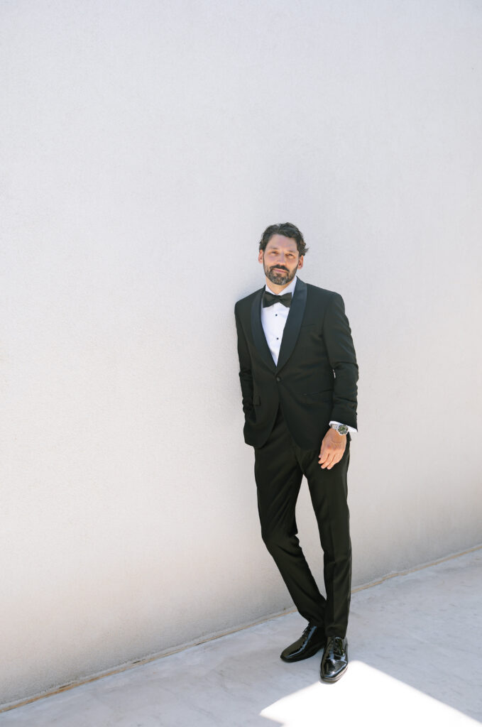 A man with dark hair and a beard wears a black tuxedo with a white shirt and black bow tie, standing against a plain light-colored wall and looking relaxed with one hand in his pocket.