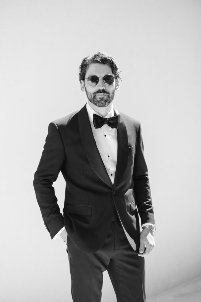 A man with a beard and mustache wearing a black tuxedo, bow tie, and sunglasses stands confidently against a plain light background. He has one hand in his pocket and a slight smile. The image is in black and white.