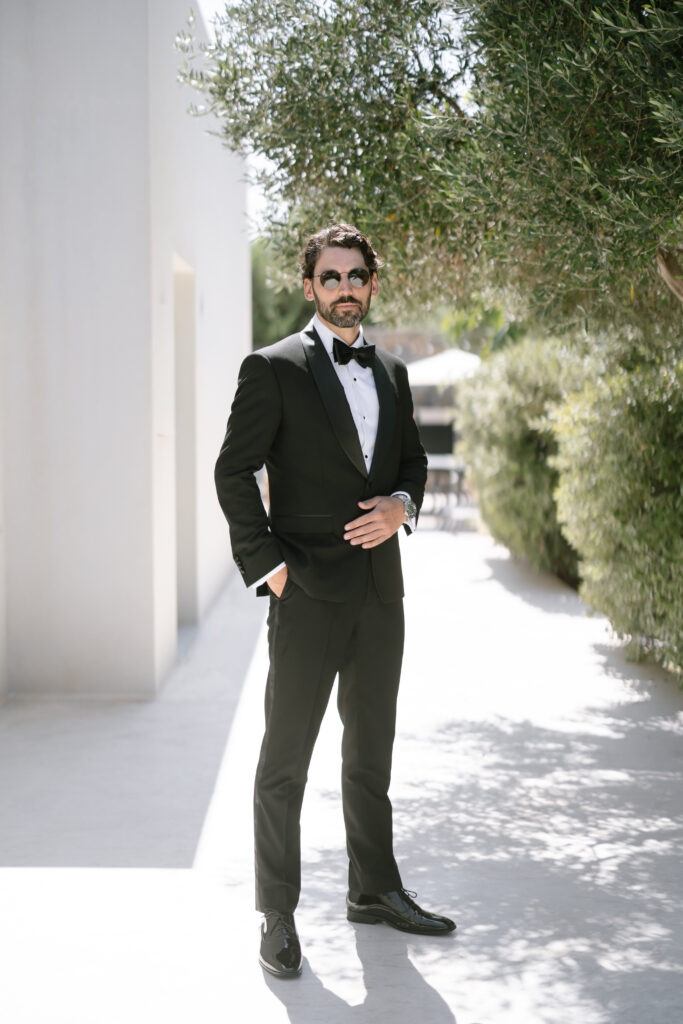A man wearing a black tuxedo, bow tie, and sunglasses stands confidently outdoors on a sunny day, with greenery and a white building in the background.
