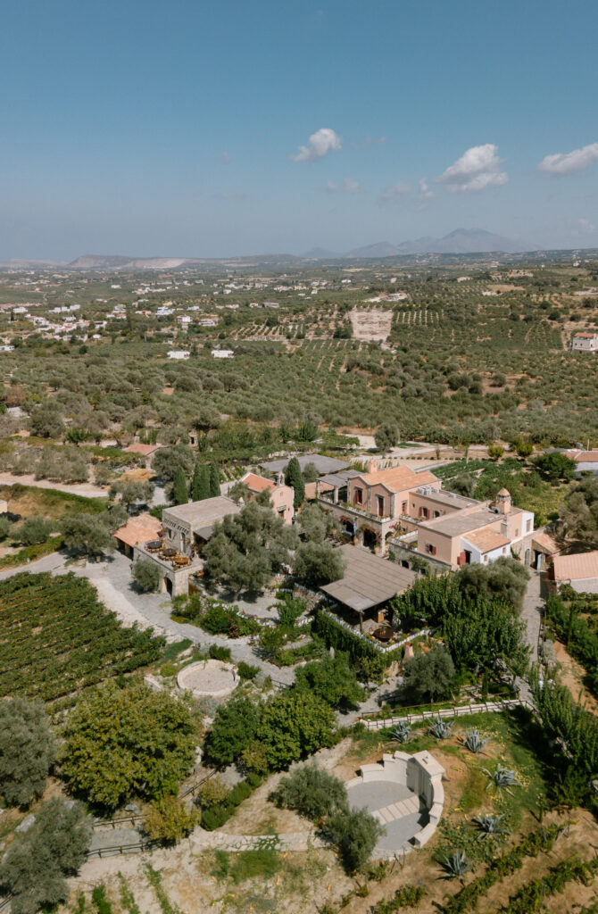 Aerial view of a Mediterranean countryside estate with several rustic buildings surrounded by olive trees, gardens, and fields, with rolling hills and distant mountains under a blue sky with scattered clouds.