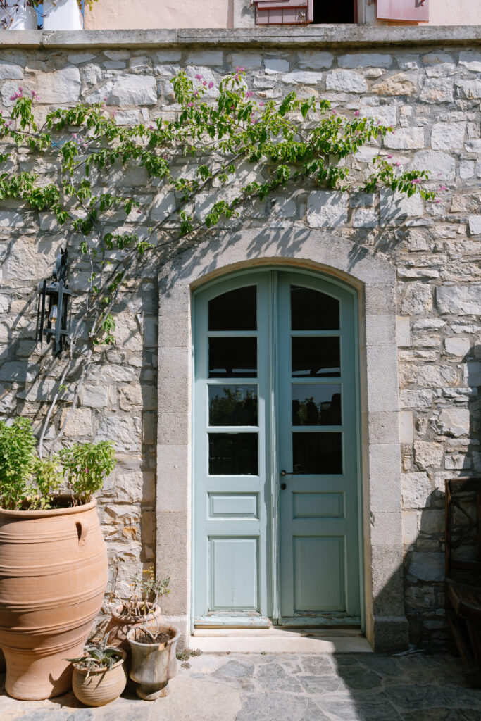 A light blue double door set in a stone wall, with green vines above and large terracotta pots with plants beside the entrance, bathed in natural sunlight.