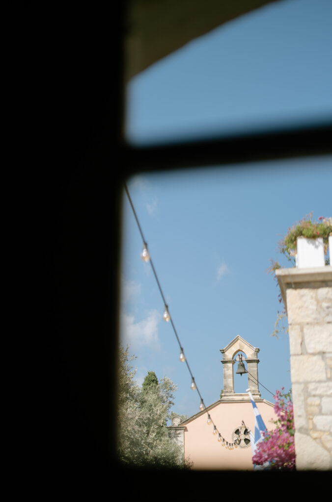 A view through a window of a small church with a bell tower, string lights, and blue sky. Part of a stone building with flowers is visible on the right side.