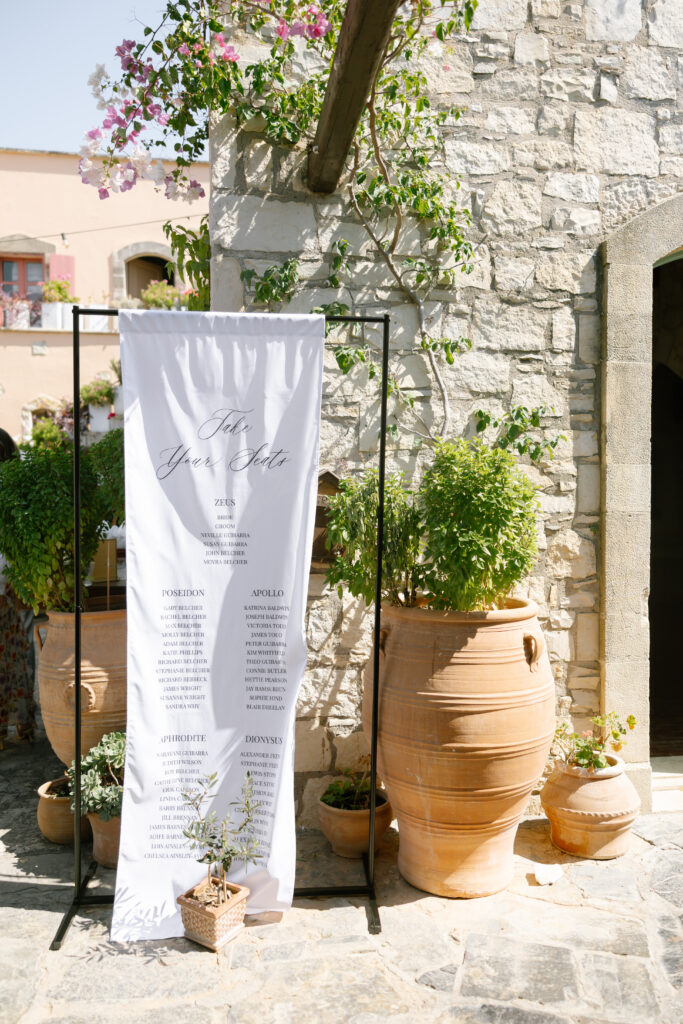 A decorative seating chart on white fabric hangs on a black metal stand next to large terracotta pots with green plants, set against a rustic stone wall in bright sunlight.