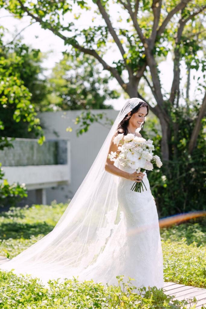 A bride in a white lace gown and long veil walks outdoors holding a bouquet of white flowers, surrounded by lush greenery and sunlight filtering through the trees.
