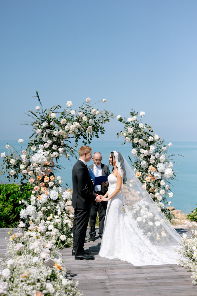 A bride and groom stand holding hands at an outdoor wedding ceremony, facing each other beneath a floral arch, with an officiant and the ocean in the background under a clear blue sky.