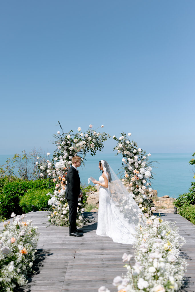 A bride and groom stand facing each other at an outdoor wedding ceremony by the sea, surrounded by floral arrangements and an arch, under a clear blue sky—captured perfectly by a Thailand wedding photographer.