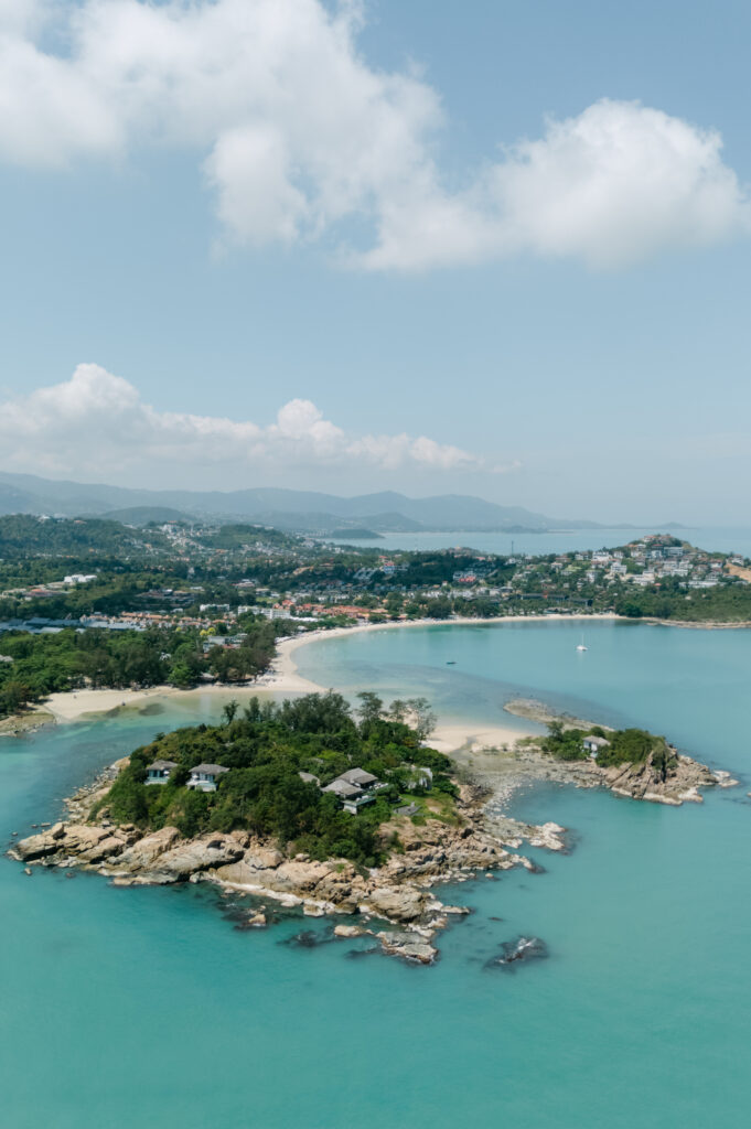 A small, lush island with rocky shores sits in turquoise water near a sandy coastline, with hills, scattered houses, and distant mountains under a partly cloudy sky.