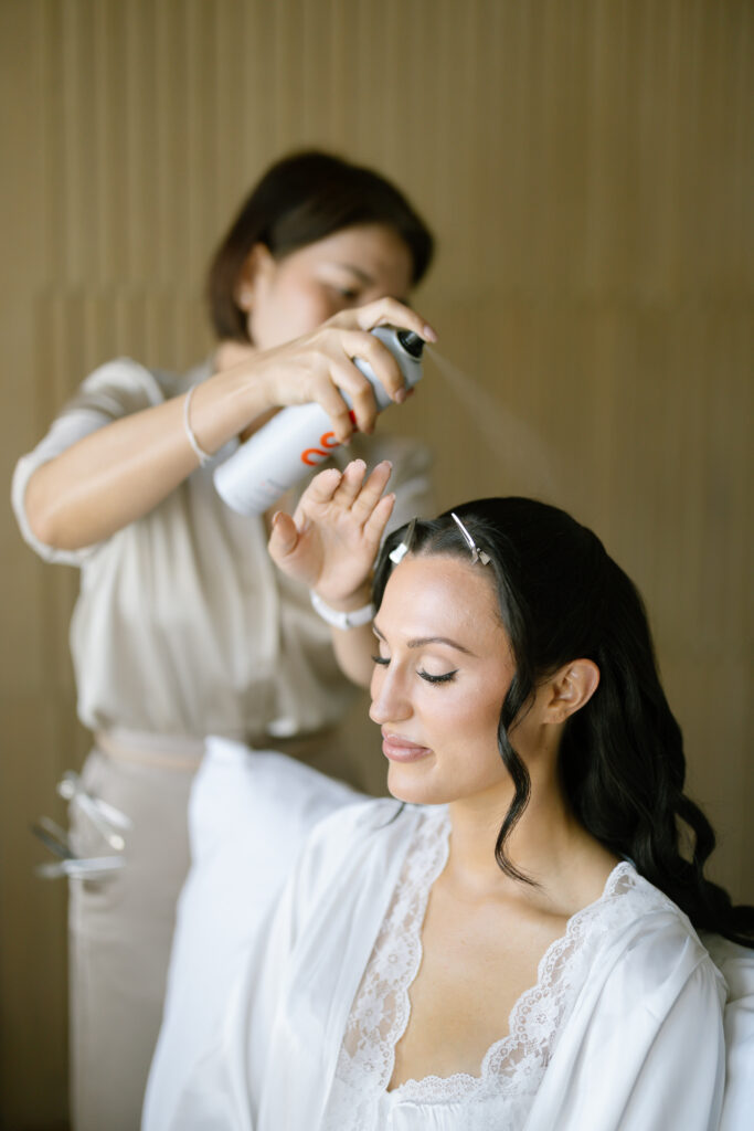 A woman in a white robe sits with her eyes closed while another person styles her hair and sprays hairspray. The setting appears calm and indoors, likely during preparations for a special event.
