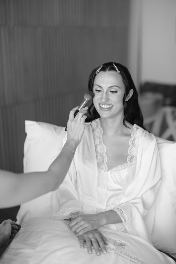 A smiling bride in a white robe sits on a bed while someone applies makeup to her face with a brush. The setting appears calm and intimate, capturing a wedding preparation moment.