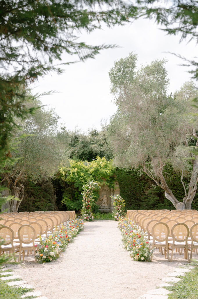 An outdoor wedding ceremony setup at Bastide Du Roy with rows of circular-backed chairs and colorful flowers lining a central aisle, ending at a floral arch surrounded by lush greenery and tall trees.