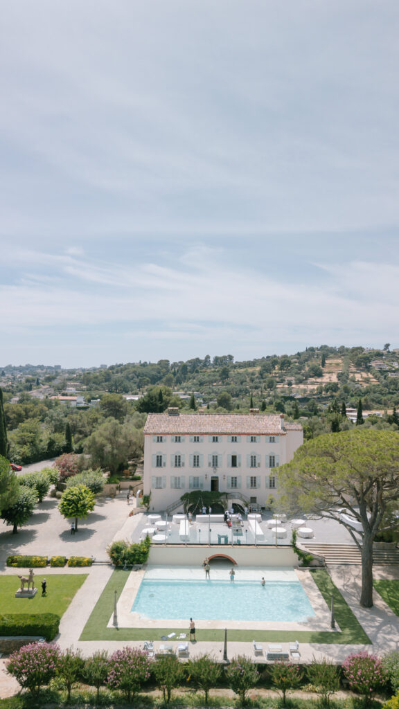 A white villa with a red-tiled roof stands behind a rectangular outdoor pool, surrounded by greenery and landscaped gardens; rolling hills and trees are visible in the background under a partly cloudy sky. Drone shot of Bastide Du Roy
