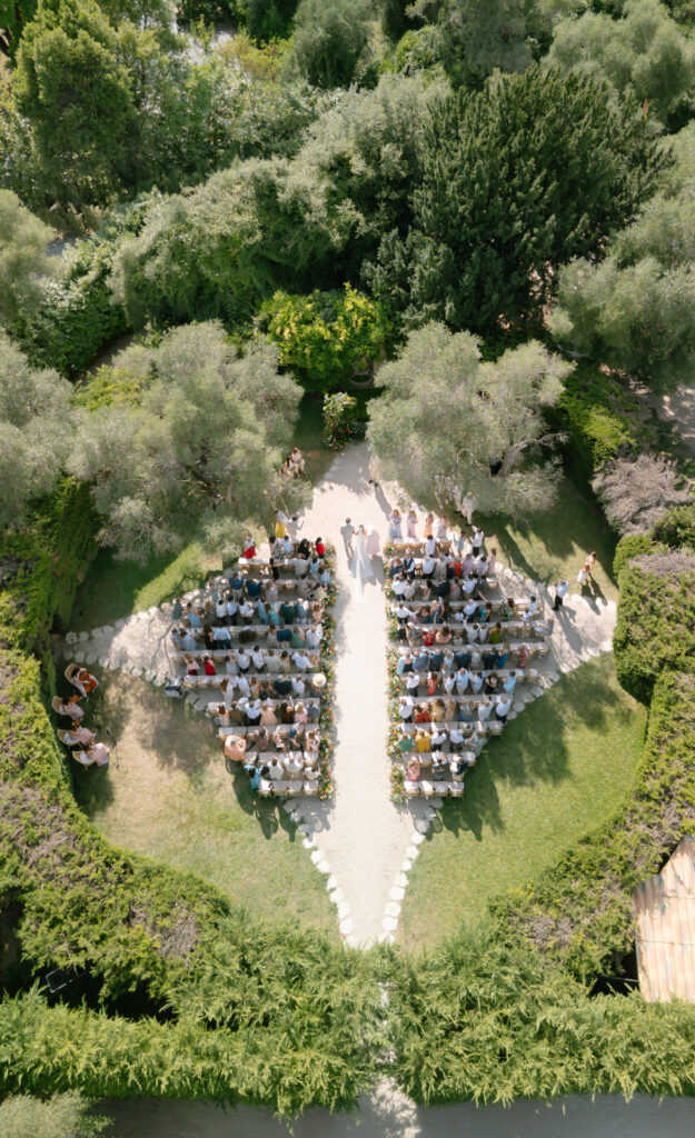 Aerial view of an outdoor wedding ceremony at Bastide Du Roy, with guests seated in two sections forming a leaf-like shape, surrounded by lush green gardens and trees. The couple stands at the front, facing the guests.