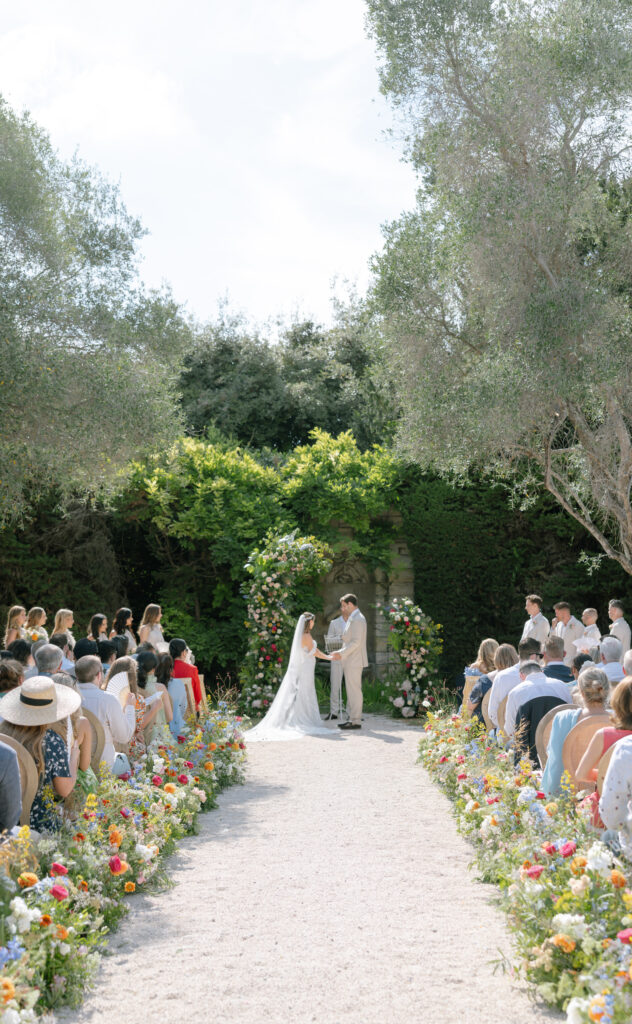 A bride and groom stand at an outdoor altar surrounded by lush greenery and colorful flowers, holding hands during their wedding ceremony as guests watch from both sides of a flower-lined aisle.
