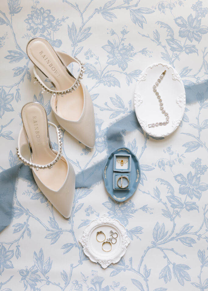 A pair of elegant white heels with pearl straps, a pearl necklace, gold rings, and a gold ring with a blue stone are displayed on ornate white and blue trays atop a light blue floral-patterned surface.