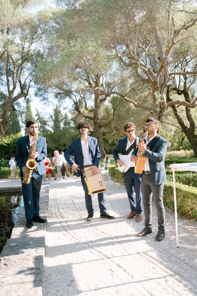 Four musicians in blue suits perform outdoors on a sunny day, playing saxophone, guitar, and percussion, surrounded by trees and greenery. People are visible in the background.