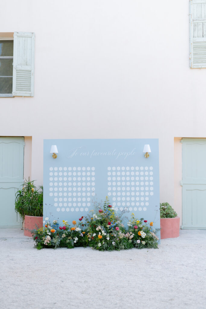 A pastel blue seating chart board with white circles and the phrase “To our favorite people” stands against a pale building, decorated with fresh colorful flowers and greenery at the base.