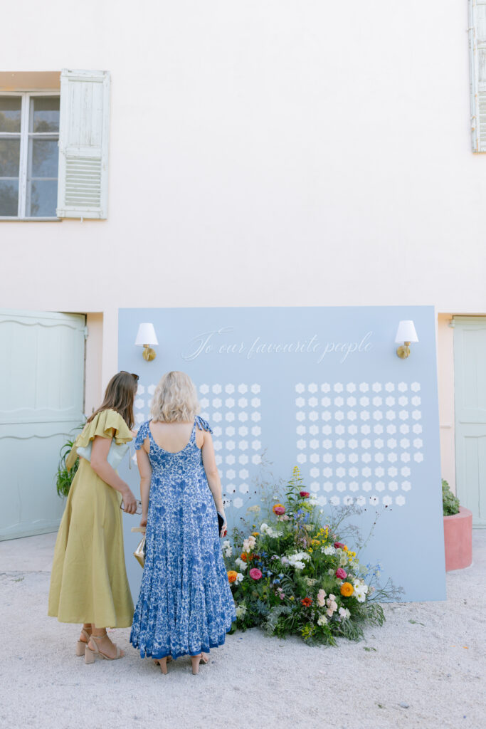 Two women in colorful dresses stand in front of a pastel blue seating chart decorated with white cards and floral arrangements, against a pale wall with windows and mint green doors. The sign reads, “To our favourite people.”.