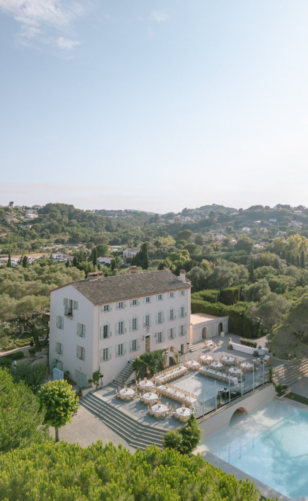 Elegant chateau with light facade, surrounded by lush greenery, set for an outdoor event with round tables and chairs by a pool; hills and houses visible in the background under a clear sky.