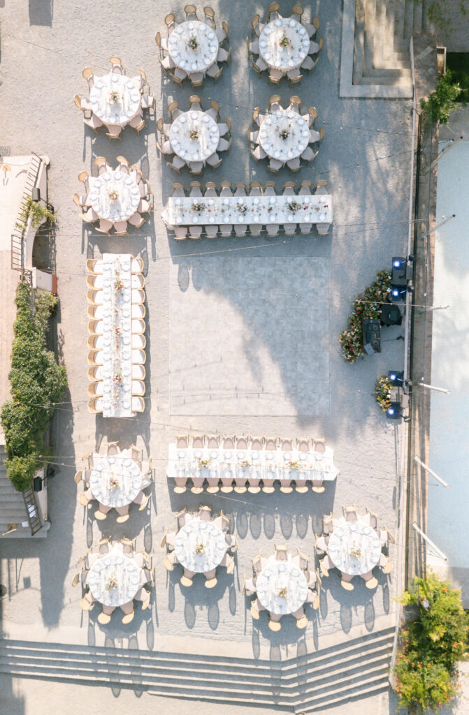 Aerial view of an outdoor event setup with round and rectangular tables arranged neatly, each set with white tablecloths and chairs, surrounding an open square space on a stone courtyard.