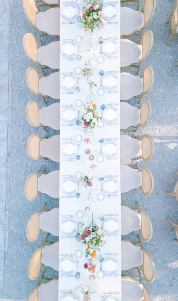Overhead view of an elegant outdoor dining table set for a formal event, with beige chairs, white tablecloth, plates, glasses, and colorful floral centerpieces arranged along the middle.