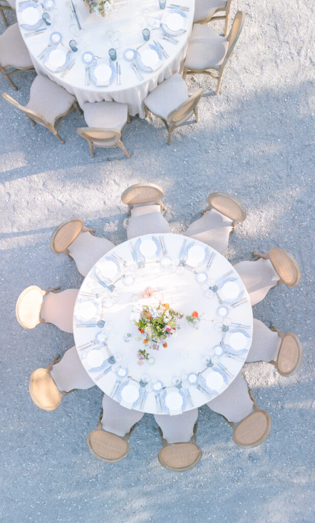 Aerial view of two round banquet tables set outdoors with white tablecloths, place settings, wooden chairs, and floral centerpieces, arranged on a light gravel surface.