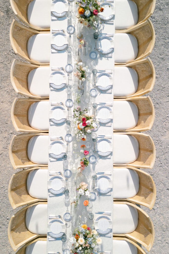 Aerial view of an outdoor dining table set for a formal event, with elegant place settings, glassware, and colorful floral centerpieces surrounded by beige cushioned chairs.