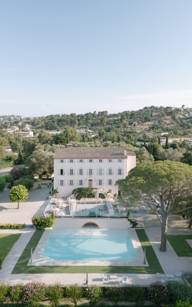 A large white villa with a tiled roof stands behind a rectangular swimming pool, surrounded by greenery and gardens, with hills and trees in the background under a clear blue sky.