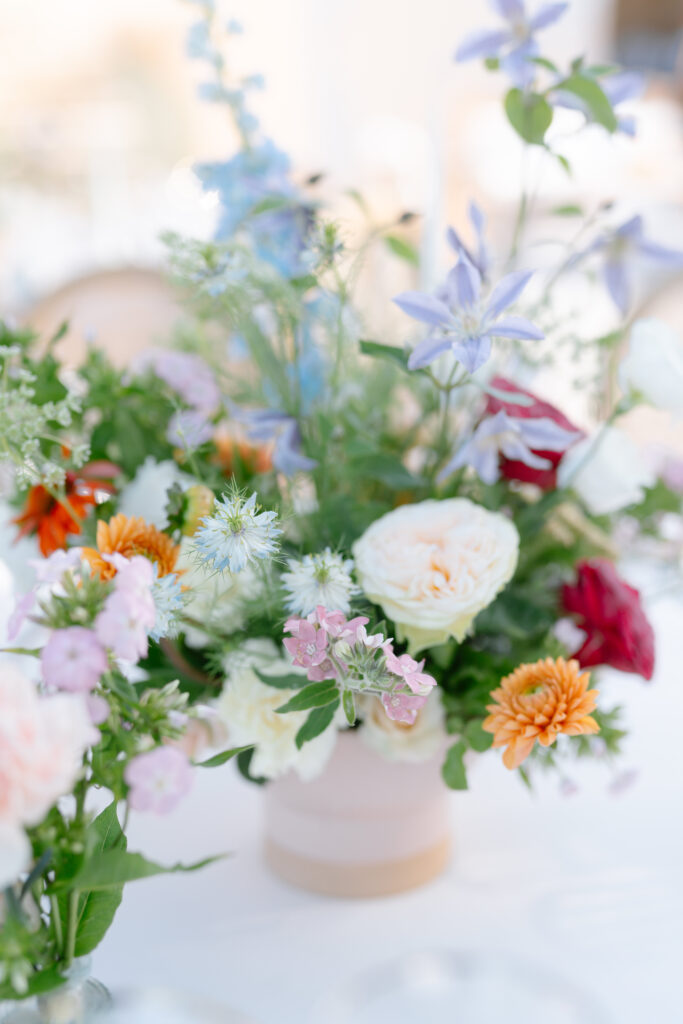 A pastel floral arrangement sits on a white table, featuring roses, chrysanthemums, and delicate wildflowers in soft pinks, purples, oranges, and whites, with lush green foliage.