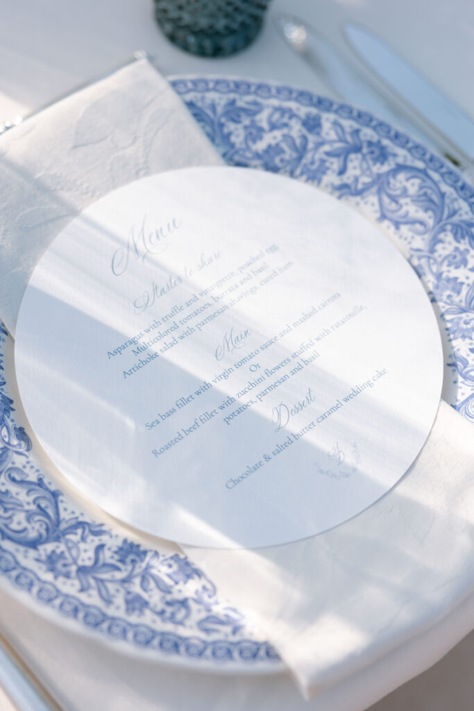 A round, white wedding menu card with elegant script lies on a white napkin atop a blue and white patterned plate, set on a formal table with sunlight casting soft shadows across the scene.
