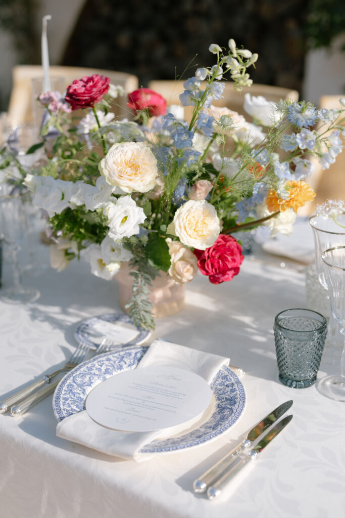 A beautifully set table with a white tablecloth, blue and white patterned plate, menu, silver cutlery, clear glass, and a floral centerpiece featuring pink, white, and yellow flowers in soft sunlight.