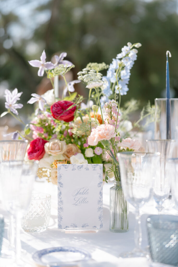 A bright, elegant table setting outdoors featuring a floral arrangement with pink, red, and blue flowers, clear glassware, and a decorative card labeled Table Lillie in a blue floral border.