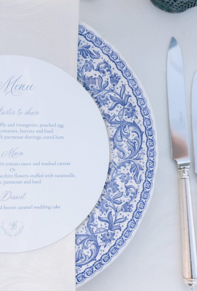 A close-up of a blue and white decorative plate with an elegant printed menu partially covering it. A silver knife and fork are placed on the right side, all set on a white tablecloth.