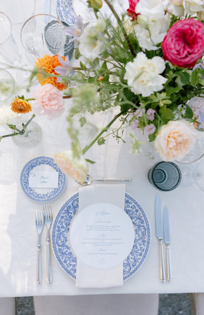 Elegant table setting with blue and white patterned plates, silver cutlery, a white napkin, a printed menu, and a large floral arrangement featuring colorful roses and greenery on a white tablecloth.