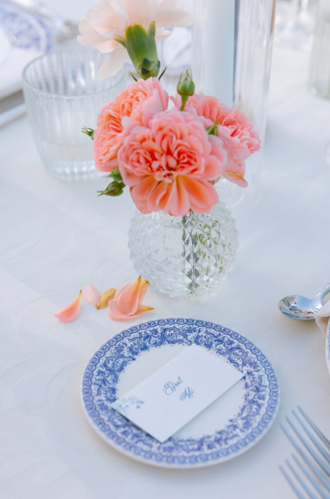 A small vase of pink flowers sits on a white tablecloth beside a decorative blue and white plate holding a place card that reads Elise. Silverware and a few pink petals are nearby.