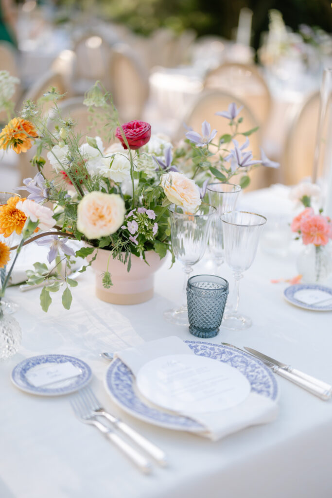A close-up of an elegant outdoor table setting with blue and white patterned plates, silver cutlery, glassware, a menu, and a pastel floral centerpiece featuring roses and greenery on a white tablecloth.