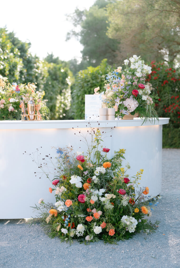 A modern white outdoor bar decorated with vibrant floral arrangements in pink, yellow, orange, and white, surrounded by greenery and bathed in soft natural light.