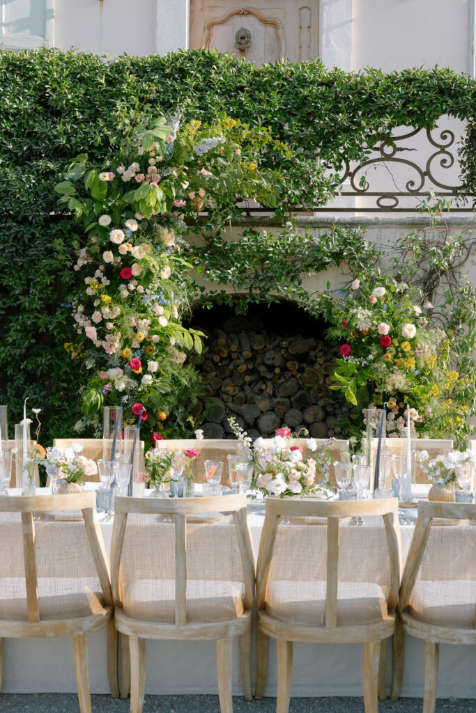 An elegant outdoor dining table is set with floral centerpieces, glassware, and wooden chairs. Behind the table, a lush green arch with pink, white, and red flowers frames a stone fireplace filled with stacked logs.