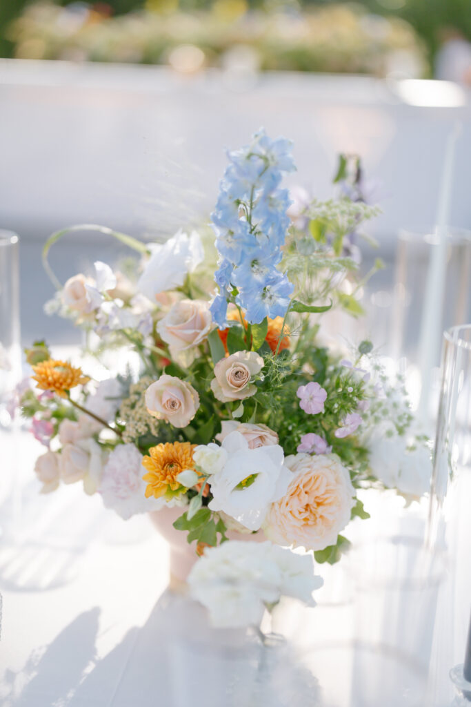 A pastel floral arrangement features cream roses, white lilies, yellow flowers, purple blooms, and a tall blue delphinium, set in a vase on a sunlit table outdoors.