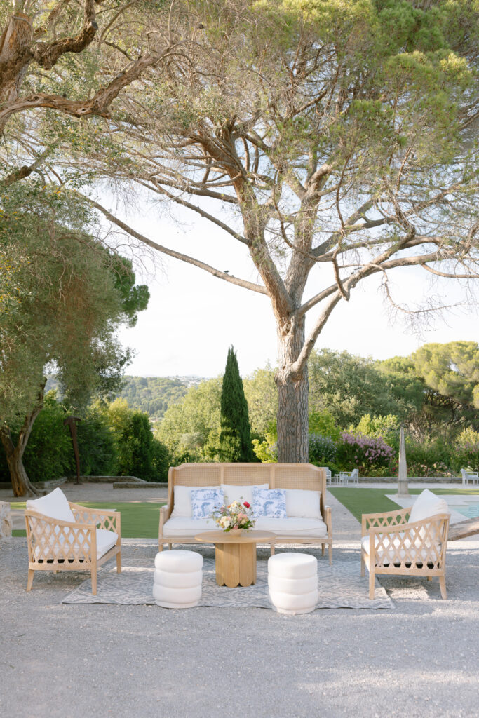 Outdoor seating area with a sofa, two armchairs, and round white ottomans arranged around a small wooden table, set under a large tree on gravel, with greenery and distant hills in the background.